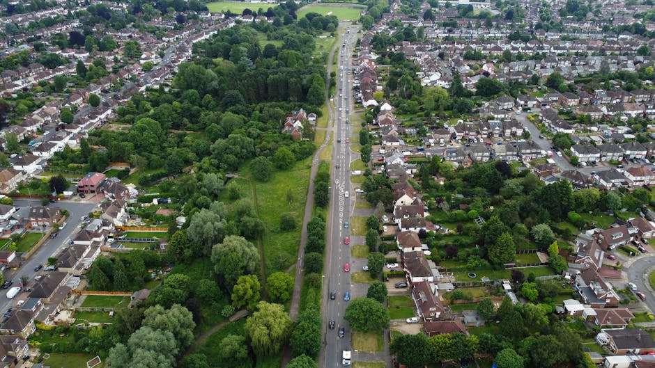 Aerial view of Colney Hatch Lane in Barnet, showing a busy street with multiple lanes of traffic including cars and vans moving in both directions, flanked by residential houses with driveways, gardens, and trees. To the left of the road, there is a green park area with lush trees, grass, and pathways. On the right side, there are terraced and semi-detached houses with front gardens, some containing vehicles and garden furniture. The street appears to be part of a well-established suburban neighbourhood, with clear signage and a mix of greenery and built environment, illustrating the typical setting for home relocations and furniture transport activities. This scene is relevant to house removals and movement logistics, as provided by Man with Van Colney Hatch, especially in the context of applying for Barnet Council permits for removals on Colney Hatch Lane.
