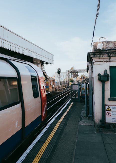 A modern train parked alongside a train platform at New Southgate Station in Colney Hatch. The train has a sleek, streamlined design with beige and red exterior colors, and tinted window panels. The platform features safety yellow tactile paving along the edge for passenger safety, with a green warning sign on the adjacent small white building, which houses electrical or communication equipment. Nearby, a set of stairs and a metal railing lead towards the station entrance, while in the background, railway tracks extend into the distance, intersected by overhead electrical catenary wires supported by metal poles. The sky above is partly cloudy with soft natural light illuminating the scene, providing a clear view of the station's infrastructure, ideal for reference in house removals or moving logistics, as conducted by a company like Man with Van Colney Hatch during furniture transport or home relocation processes.