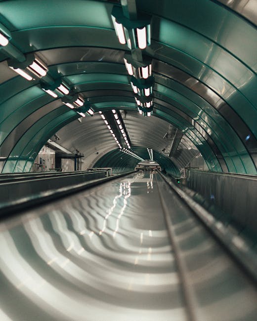 A downward-looking view of an underground urban transit tunnel with smooth, curved metallic walls illuminated by linear fluorescent lights on the ceiling, leading into a distant tunnel entrance. The shiny metal surface of the moving walkway in the foreground reflects the overhead lighting, and the tunnel's interior is designed to support train or metro transport. This setting is related to infrastructure supporting house removals and relocation services near New Southgate Station, as seen in the context of services offered by Man with Van Colney Hatch, which includes handling logistics for furniture transport and moving processes.