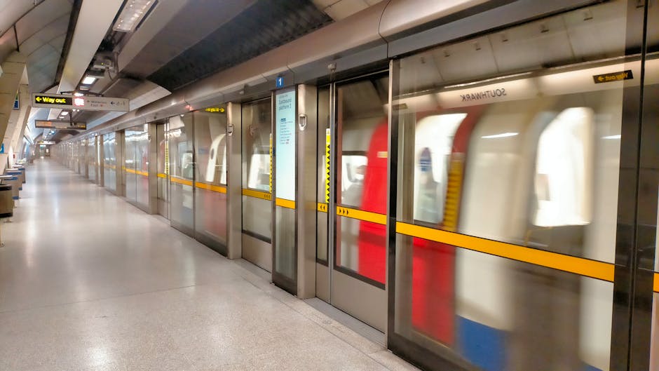 A modern train parked alongside a train platform at New Southgate Station in Colney Hatch. The train has a sleek, streamlined design with beige and red exterior colors, and tinted window panels. The platform features safety yellow tactile paving along the edge for passenger safety, with a green warning sign on the adjacent small white building, which houses electrical or communication equipment. Nearby, a set of stairs and a metal railing lead towards the station entrance, while in the background, railway tracks extend into the distance, intersected by overhead electrical catenary wires supported by metal poles. The sky above is partly cloudy with soft natural light illuminating the scene, providing a clear view of the station's infrastructure, ideal for reference in house removals or moving logistics, as conducted by a company like Man with Van Colney Hatch during furniture transport or home relocation processes.
