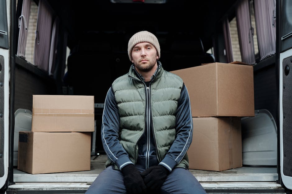 A young man with light brown hair, dressed in a blue zip-up hoodie, white t-shirt, and navy trousers, is sitting on the edge of a moving van's open rear doorway. He appears to be taking a break during