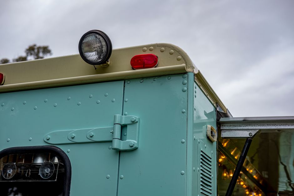 A small single-axle trailer parked on a paved driveway next to a dense green hedge, with a blue tarpaulin cover secured over its contents using black straps. The trailer has a metal frame, a single wh