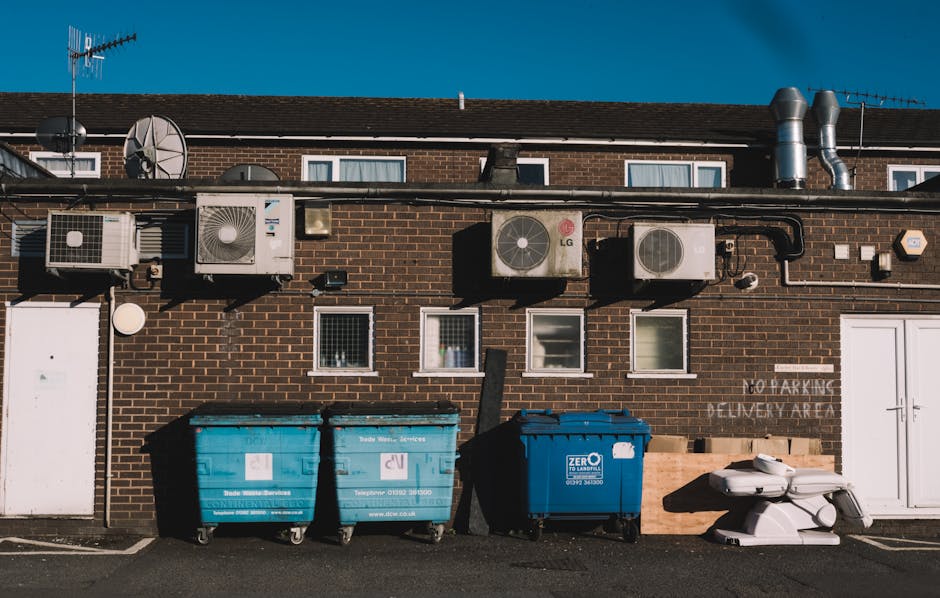 A row of three large wheeled waste disposal bins positioned on the pavement in front of a brick residential building, with two blue bins and one darker blue recycling bin, each marked with logos and contact details. Behind the bins, the building's exterior wall features multiple small windows with white frames, and various ventilation units and air conditioning systems are mounted above, connected by black piping. To the right of the bins, there are stacked white plastic waste containers and cardboard sheets. The building’s rear wall has a white door, a sign indicating no parking or delivery zones, and multiple rooftop satellite dishes and metallic ventilation ducts. The scene is well-lit by daylight, suggesting a clear day, and the environment is indicative of a typical urban residential area, possibly during a home relocation or clearance process, with Man with Van Colney Hatch providing waste removal or moving services.