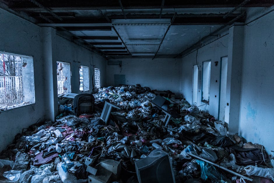 An interior room filled with a large quantity of assorted household waste and debris, including plastic bottles, bags, cardboard, and broken electronic devices, scattered across the floor. The room has damaged walls with peeling paint and multiple windows letting in natural light, revealing the cluttered environment. In the background, there are discarded furniture items and tangled cables. Outside the windows, a chain-link fence and some trees are visible. This cluttered scene illustrates a property dealing with bulky waste removal, often part of a home relocation or cleaning process, performed by Man with Van Colney Hatch to facilitate efficient furniture transport and packing for subsequent move or disposal.