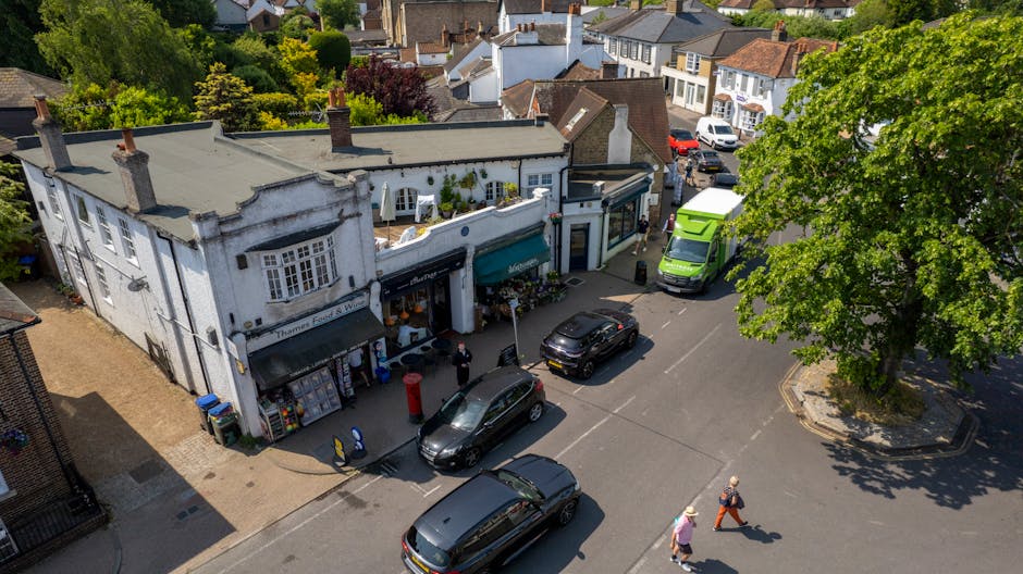 An aerial view of a street scene in Colney Hatch, showing a white, two-story mixed-use building with a shopfront on the ground floor and residential windows above, situated next to a large, leafy green tree casting shadows on the pavement. Parked along the street are several cars, including a black hatchback, a dark grey estate car, and a bright green delivery van. The sidewalk in front of the shop is occupied by a few pedestrians, some walking and others standing. The building appears to be part of a home relocation or furniture transport process, with the surrounding area demonstrating typical urban street activities. The scene is well-lit with clear daylight, showing the neighborhood's mix of commercial and residential properties, suitable for house removals and moving services. Occasionally, Man with Van Colney Hatch handles furniture transport and packing logistics within this area, as part of their house removals service.