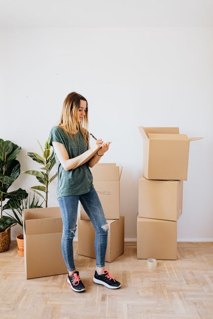 Inside a room with natural light filtering through yellow gingham curtains, several medium-sized cardboard boxes are arranged on a dark wooden surface near a bed with a grey duvet. The boxes are seale
