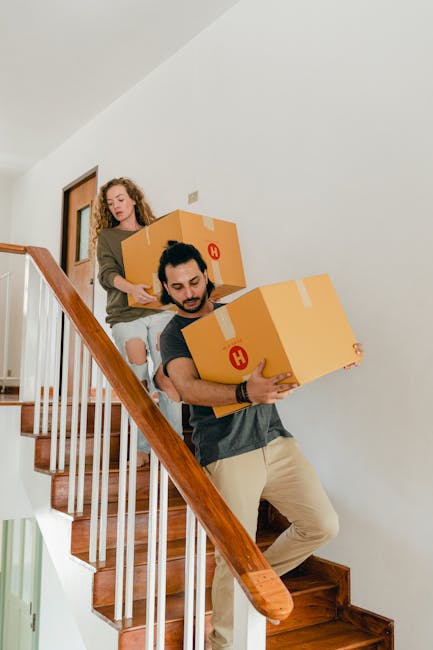 A man and a woman carrying large cardboard boxes down a wooden staircase inside a home, preparing for a house move. The man is positioned at the front, gripping a box with both hands, wearing a dark T-shirt and light-colored pants, focused on the task. Behind him, the woman is also holding a cardboard box, stepping down the stairs with a neutral expression. The staircase has wooden steps and a polished handrail, with white walls and a door visible in the background. The scene captures the process of furniture transport and packing during a home relocation, with the focus on careful handling of boxes as part of a professional moving service provided by Man with Van Colney Hatch.
