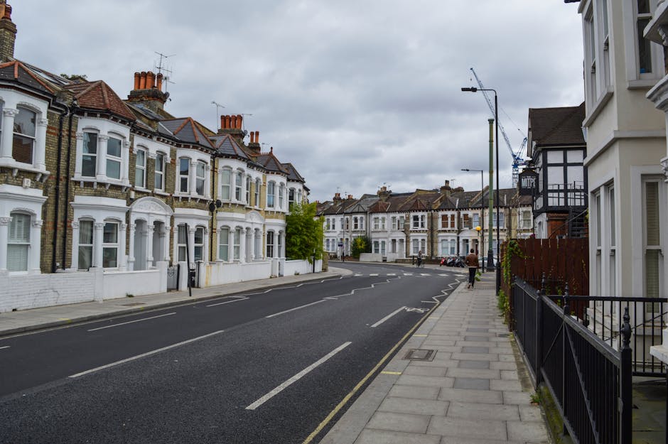 A quiet residential street in Colney Hatch featuring a row of Victorian-style terraced houses with white facades, bay windows, and tiled roofs. The pavement runs alongside the houses, with metal railings and small front gardens visible. The street is lined with tall street lamps and a few trees, under a cloudy sky. A man in casual clothing is walking along the pavement, while a large vehicle, likely a van used for home relocation, is parked nearby. Visible are cardboard boxes, moving blankets, and packing materials inside and around the vehicle, indicating an ongoing furniture transport and packing process as part of a house removal. The scene captures the urban environment and the logistical steps involved in furniture transport during home removals, with equipment such as trolleys and straps possibly used off-camera, consistent with professional removals services by Man with Van Colney Hatch.