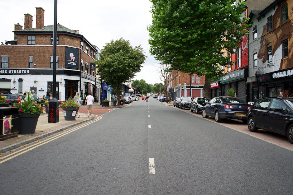 A street scene in Colney Hatch featuring parked cars lining both sides of the road and a row of commercial buildings with storefronts, one of which displays signage for a pharmacy. On the left side, there are large black potted plants with greenery and flowers, and pedestrians walking along the pavement. The street is empty of moving vehicles, with clouded sky overhead and trees providing some shade. The scene captures a typical urban environment suitable for home relocation or furniture transport, with no visible moving activity or equipment in the image, but reflecting the context of residential and commercial activity in N11 and nearby streets connected to house removals services by Man with Van Colney Hatch.